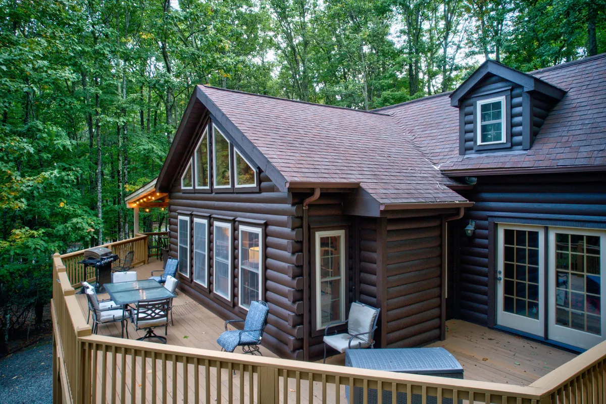 A welcoming log cabin exterior with large windows, an inviting deck furnished for outdoor dining and relaxation, surrounded by lush trees and nature. The spacious deck creates a seamless connection between indoor luxury and the tranquility of the mountain setting.