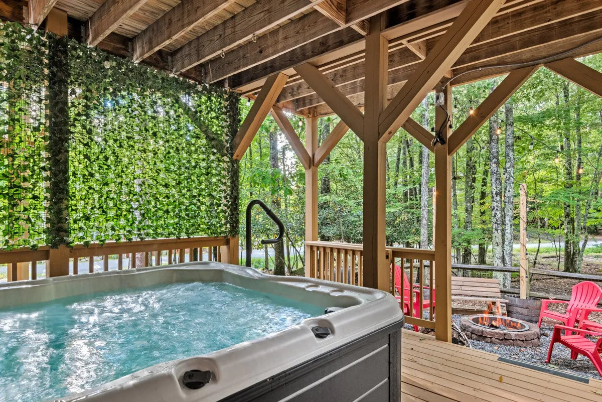 This exterior image beautifully captures a covered porch area with a bubbling hot tub, a green wall of foliage providing privacy, red Adirondack chairs, and a cozy fire pit. The space is staged for relaxation and sociable gatherings, with the natural scenery adding to the inviting atmosphere.