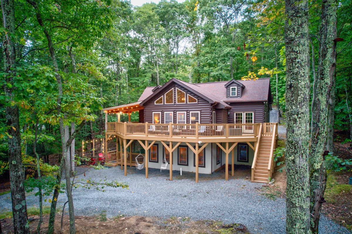 A stunning exterior shot of Mossy Rock Retreat, showcasing a spacious wooden deck with ample seating, large windows that invite natural light, and a backdrop of tall trees. The setting is tranquil, with a gravel driveway and the deck perfectly positioned for sunset views and outdoor gatherings.