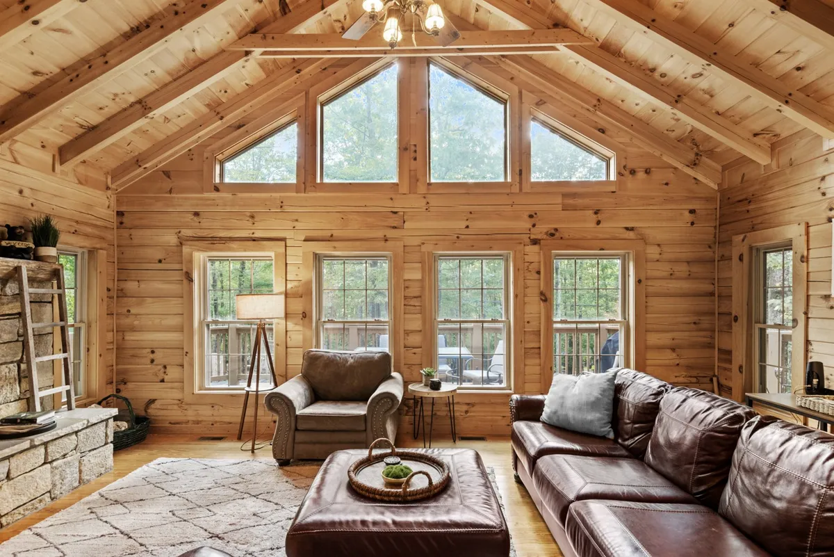 A spacious living room with soaring vaulted ceilings, exposed wooden beams, a grand stone fireplace as the focal point, and a wall of large windows that offer breathtaking views of the surrounding mountains. The room features a leather sofa and cozy décor, exuding a warm, inviting atmosphere.