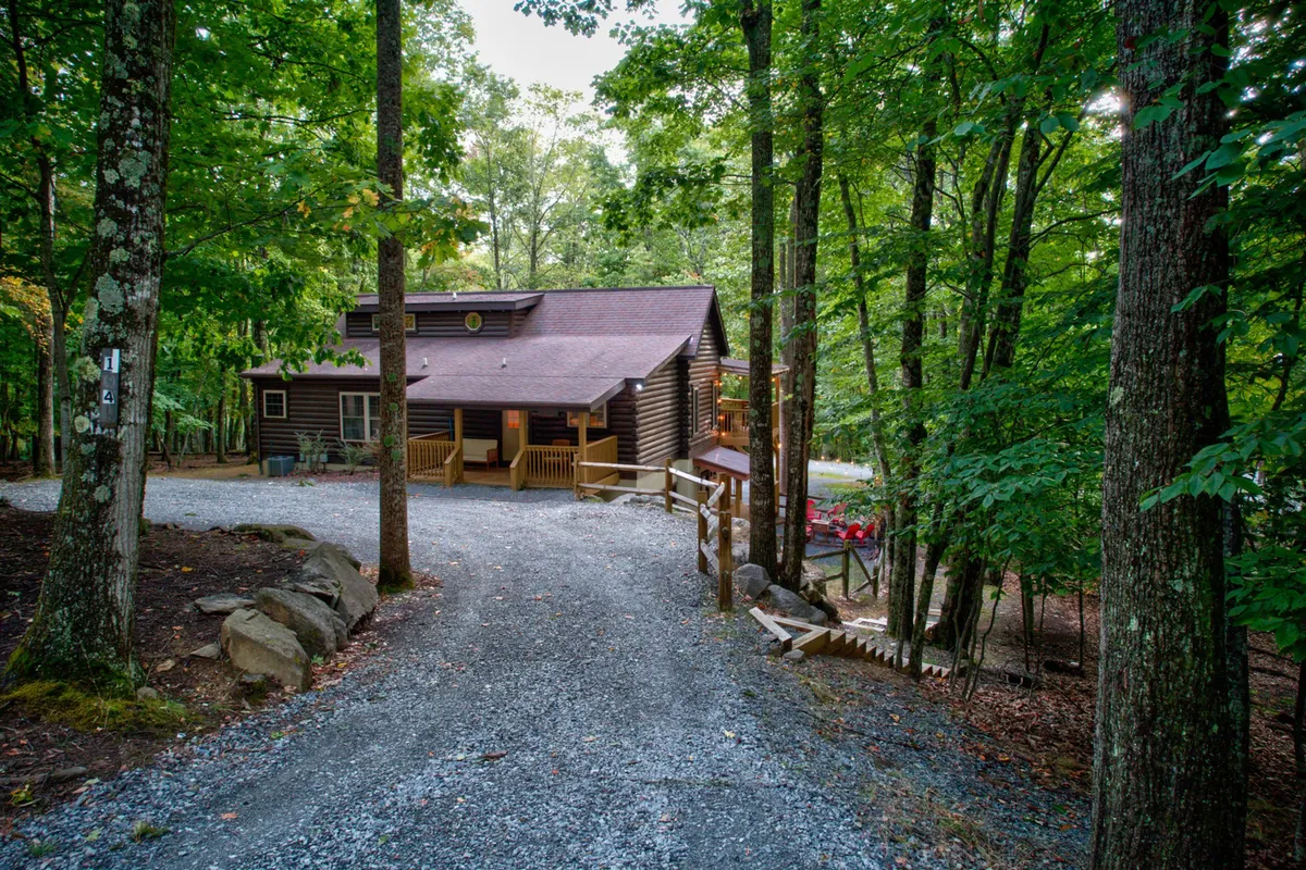 This wide-angle exterior shot depicts the log cabin nestled among tall trees, with a prominent gravel driveway, wooden deck, and inviting architecture. The tranquil surroundings hint at outdoor recreation possibilities and a peaceful mountain retreat vibe.