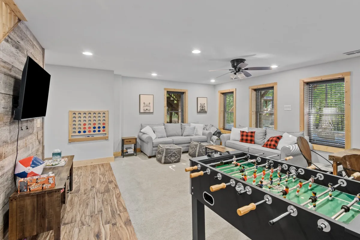 A vibrant, casual living area featuring a foosball table in the foreground, a sectional sofa for lounging, a wooden accent table, and a wall-mounted TV, suggesting a fun and interactive environment for games and relaxation.