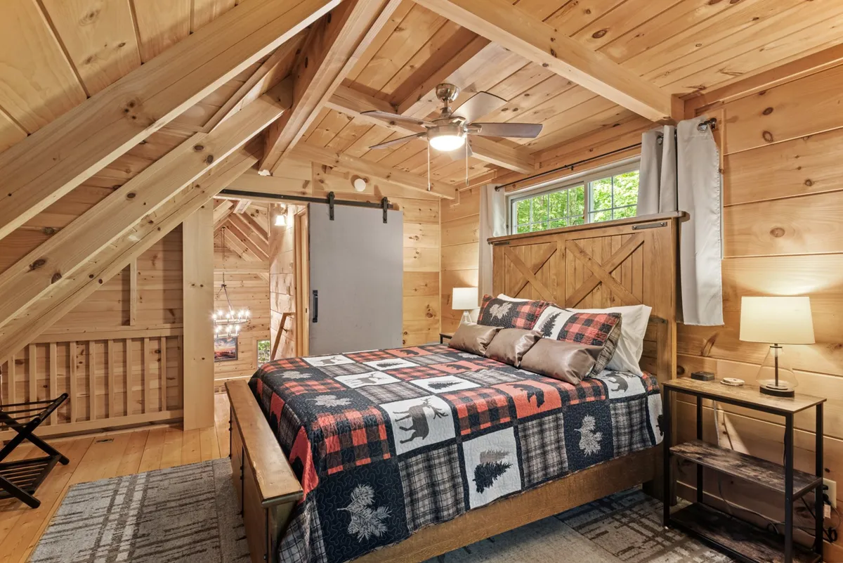 A warmly lit bedroom featuring a bed with a patterned quilt, rustic wooden walls and ceiling, and a window allowing natural light to pour in. The tranquil atmosphere is complemented by simple, cozy furnishings, creating a restful retreat.