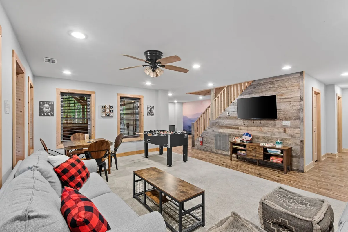A cozy entertainment room with a foosball table situated near a comfy sofa with plaid cushions. A wooden coffee table sits between the seating and a large TV mounted on the wall, creating a perfect setup for games, movie nights, and socializing.