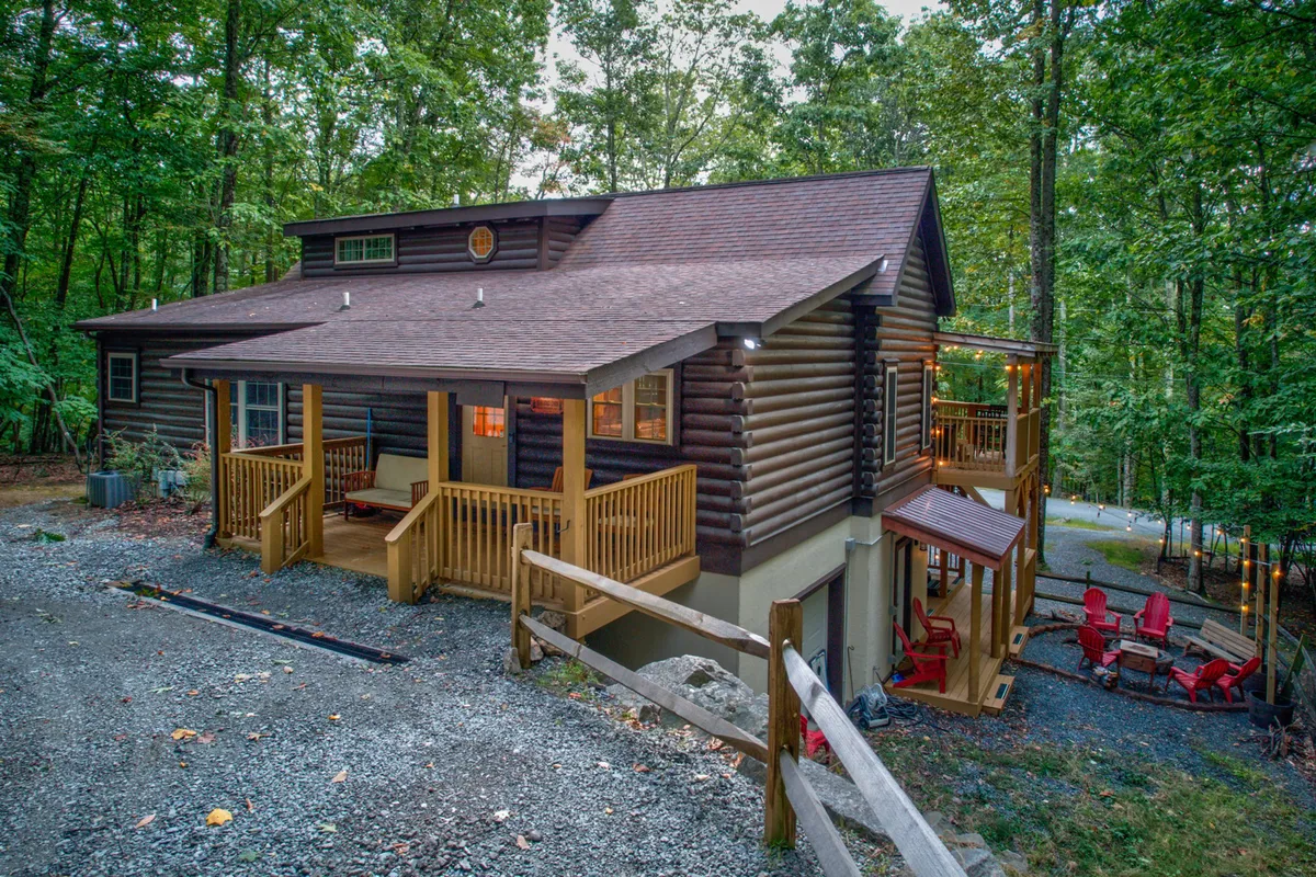 This exterior image features the log cabin's front porch with red Adirondack chairs and a well-defined fire pit area surrounded by natural greenery. The inviting setup suggests a perfect spot for gathering with friends or family for evening conversations and stargazing. The gravel driveway and ample outdoor space are also visible, connecting to the group's arrival and shared outdoor experiences.