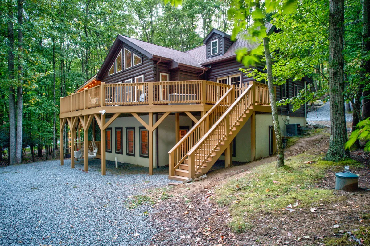 A wide view of the cabin's large wooden deck, sturdy staircase, and impressive log cabin facade, set among mature trees. The deck is furnished for outdoor gatherings, while the surrounding forest hints at nearby hiking and biking trails.