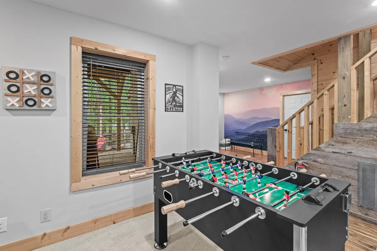 An inviting interior entertainment room featuring a foosball table, window with blinds, wooden staircase, and wall art. The room has a playful and social atmosphere, ideal for family game nights and relaxation after outdoor activities.